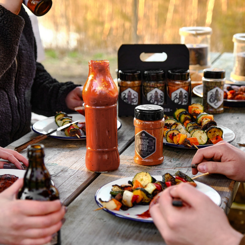 Gewürzstreuer mit Magic Dust Rub auf einem Holztisch neben Tellern mit Grillgut.