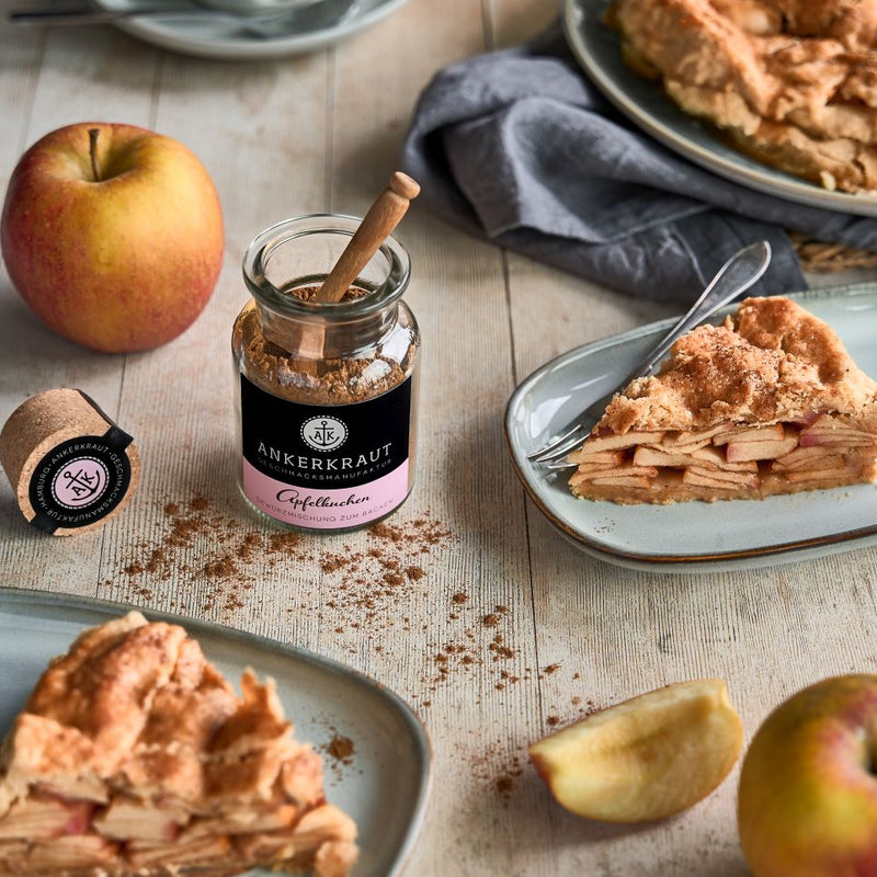 Apfelkuchen mit Zimt auf Teller, daneben Gewürzglas, Apfel und Krümel auf hellem Tisch.