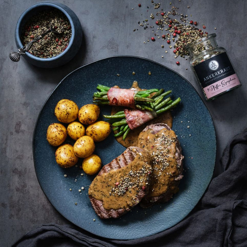 Pfeffersteak mit Kartoffeln und Bohnen im Speckmantel auf einem Teller angerichtet.