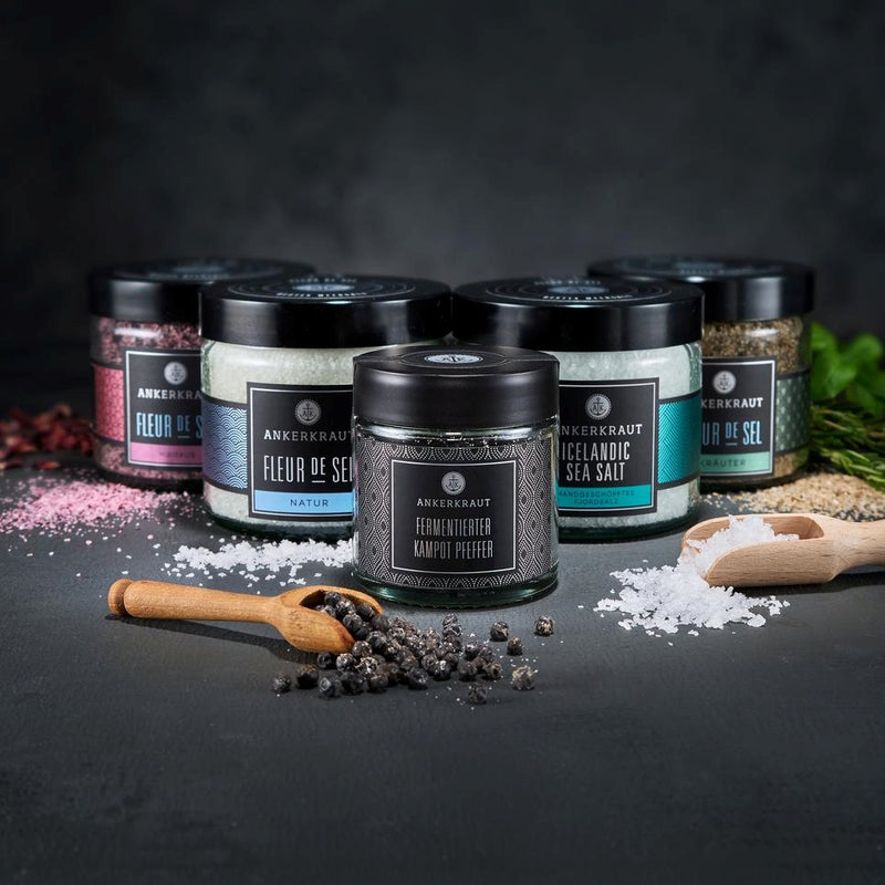 Gourmet Set mit hölzernen Gewürzlöffeln voller Fleur de Sel und Preffer vor schwarzem Hintergrund.